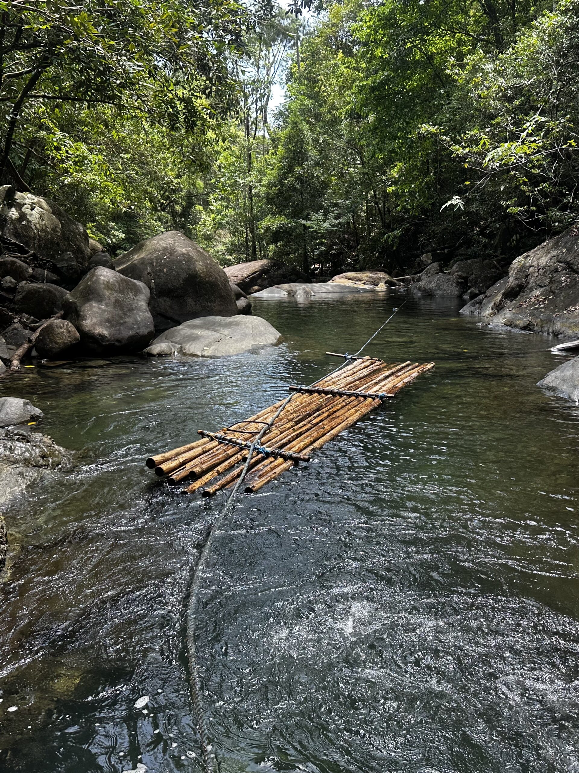 A bamboo raft tethered in a clear jungle river, boulders and rainforest canopy on both banks