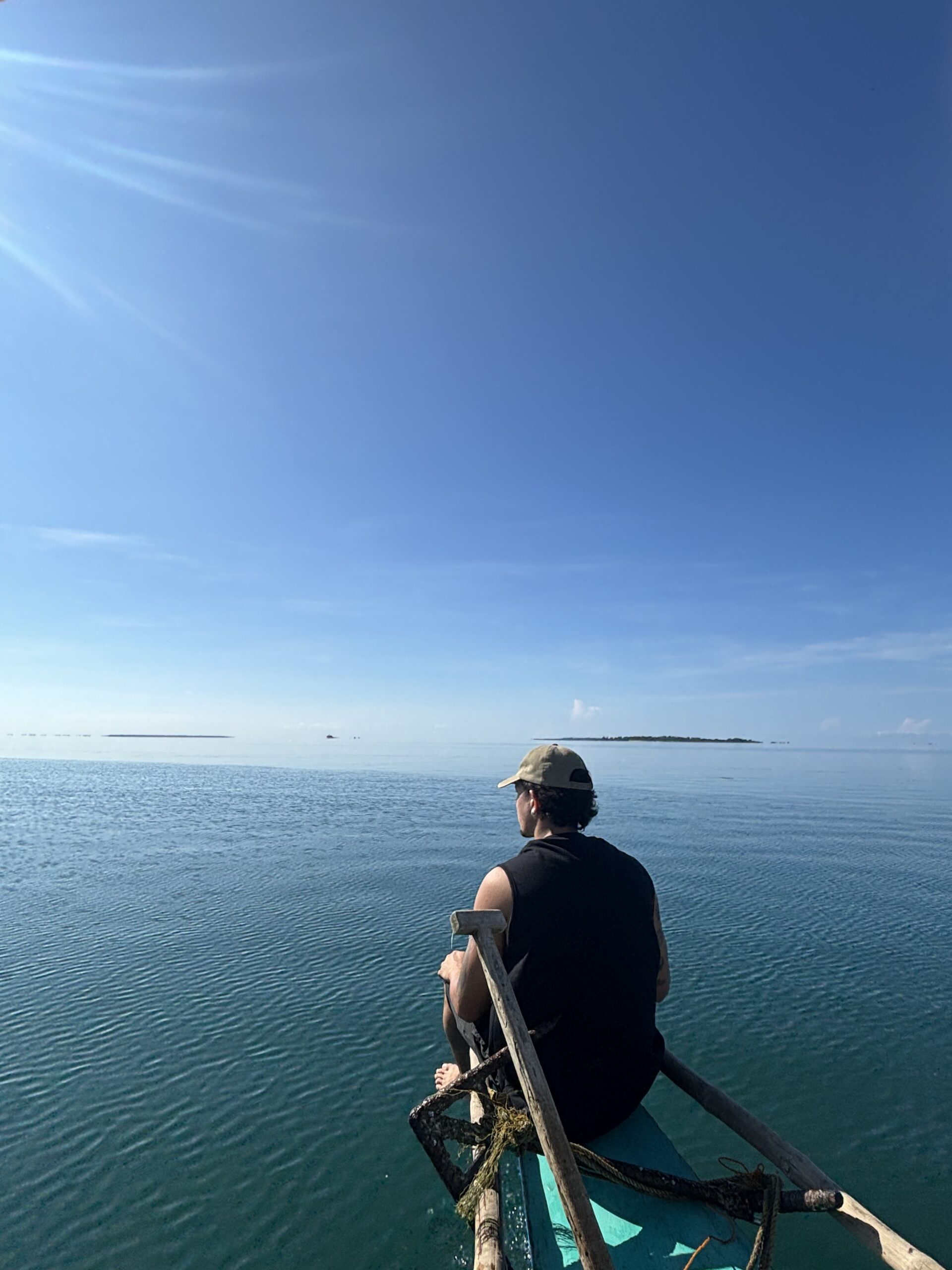 Leon sitting on the outrigger of a traditional bangka, looking out across calm turquoise water toward a distant island