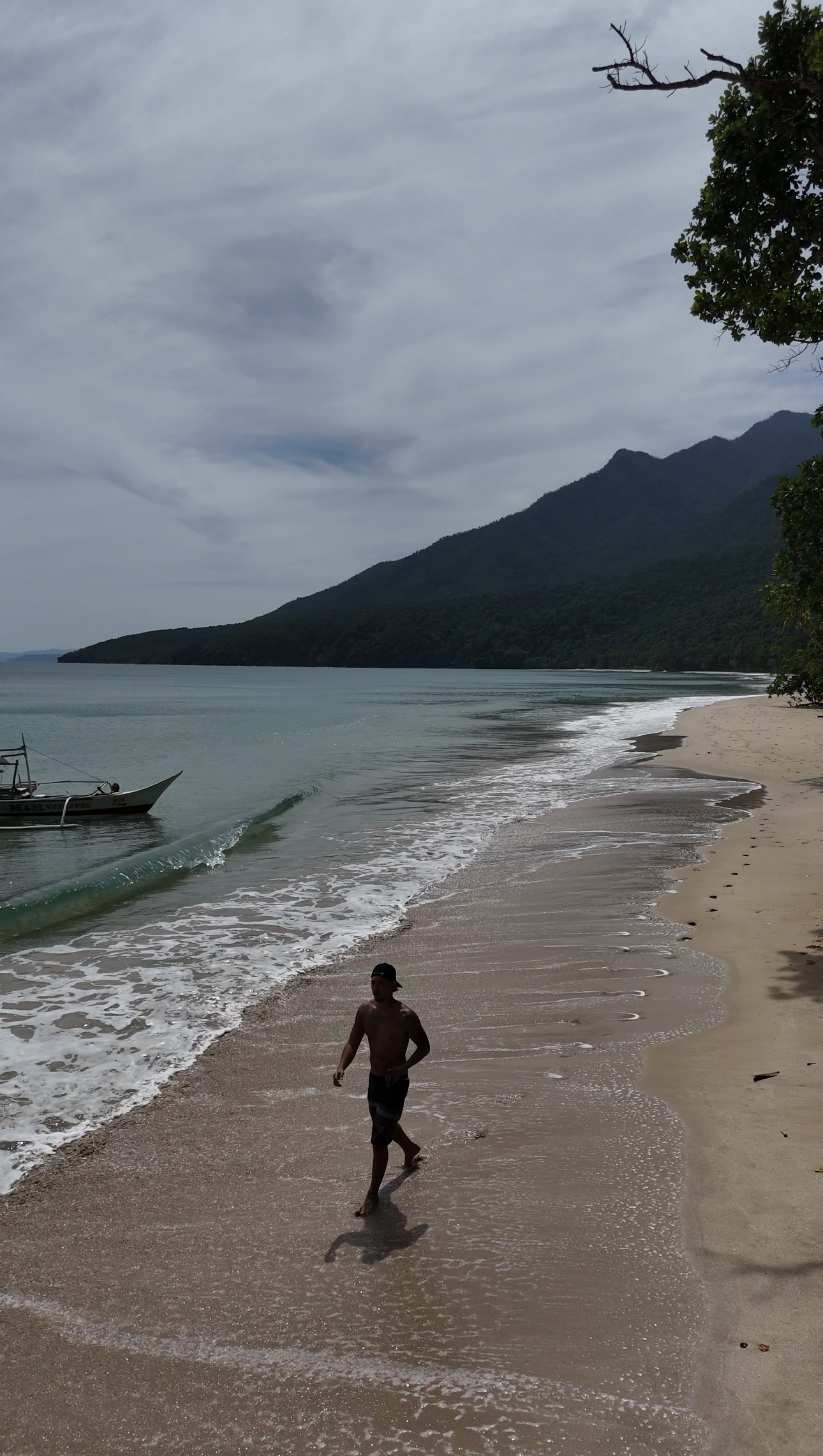 A lone figure walking the shoreline of an empty Palawan beach, forested mountains rising behind the bay